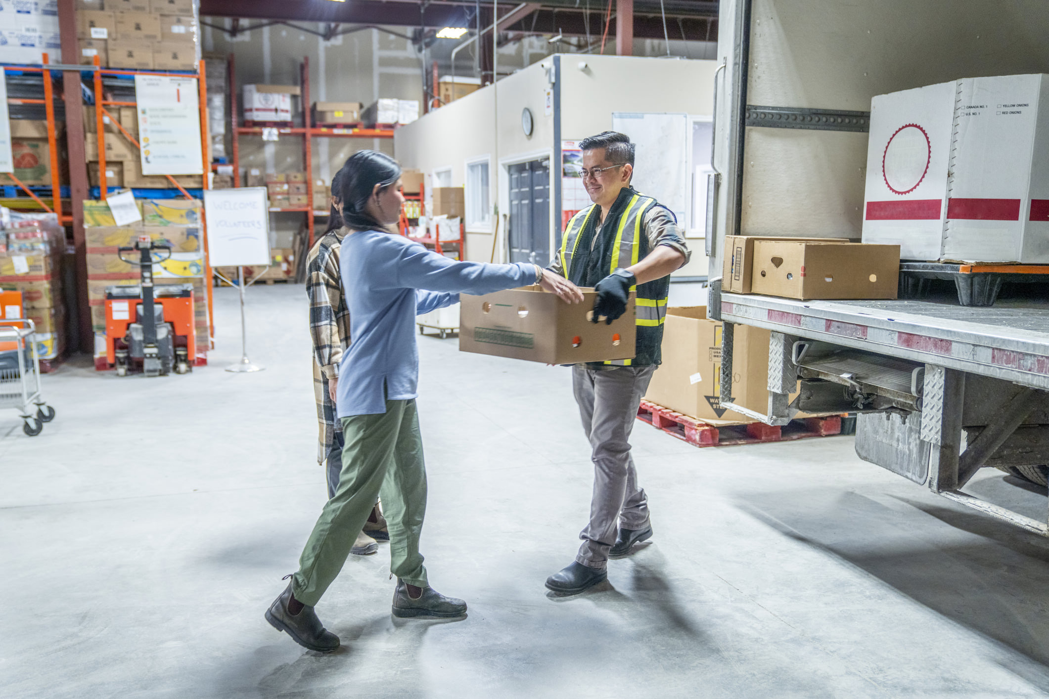 A young girl and her father are seen working together to unload a truck full of food boxes as they volunteer their time.  The father is wearing a reflective vest for safety and is passing the box to his daughter as they work together at the local Food Bank.