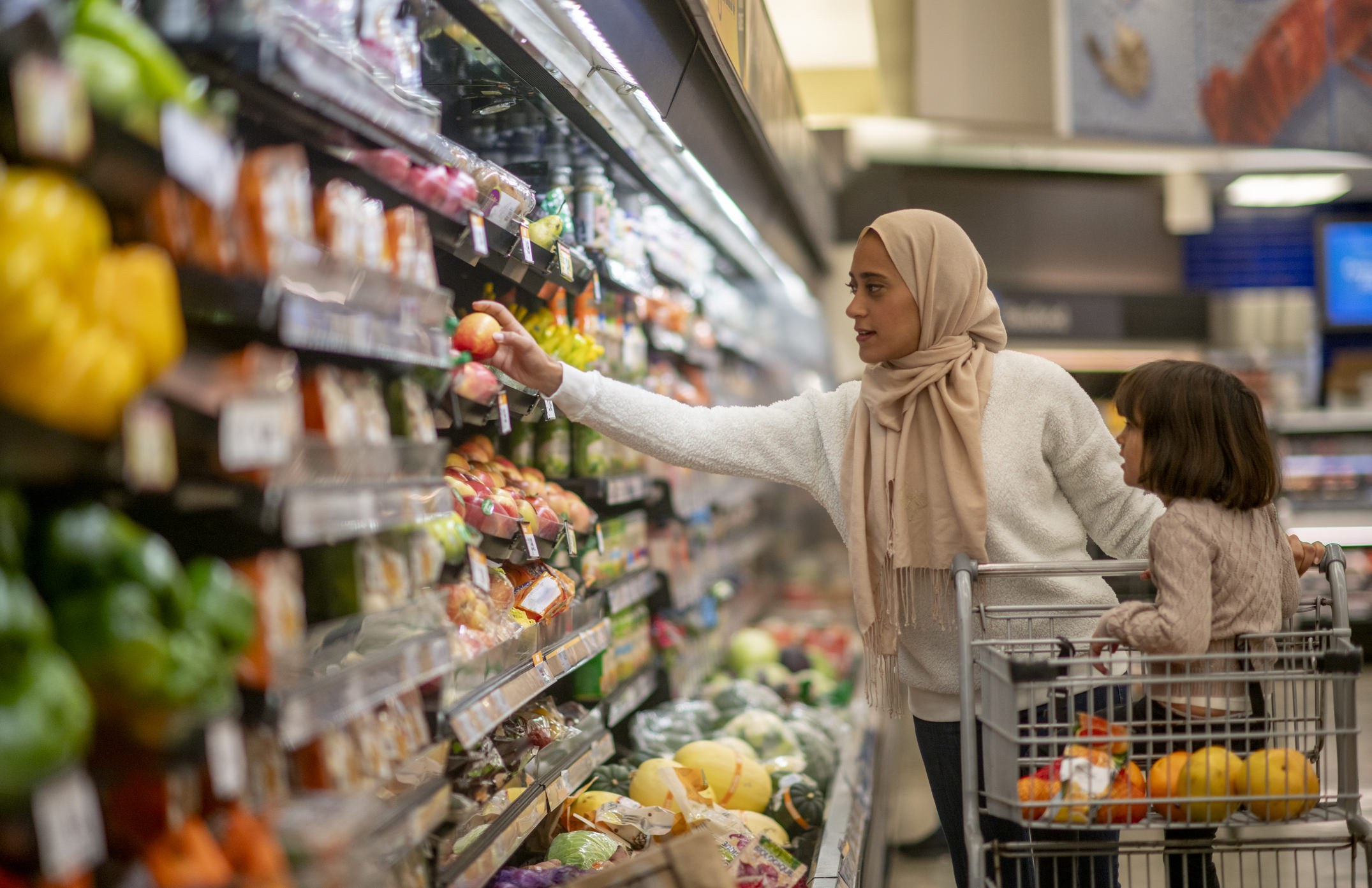 A Muslim mother wearing a hijab selects fresh produce from a supermarket shelf while her young child sits in a shopping cart, capturing everyday family shopping, warmth, and togetherness.